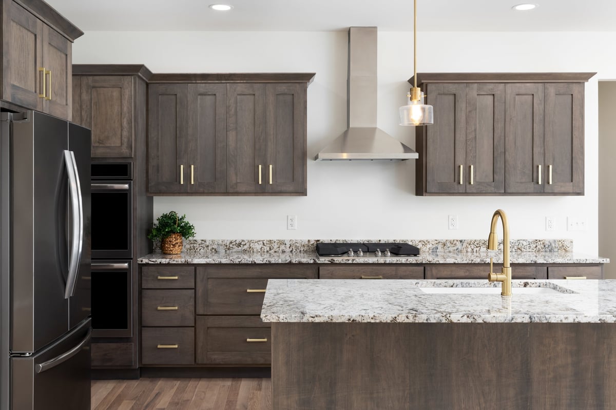 A kitchen detail with a gold faucet on a granite countertop, dark wood cabinets, stainless steel appliances, and gold pendant lights hanging above the island
